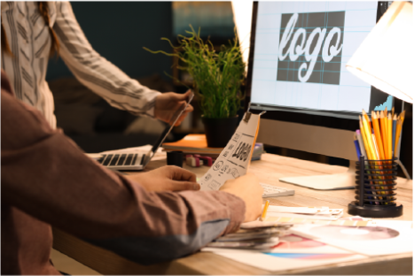 On voit deux personnes travaillant sur la conception d'un logo. Ils sont sur un bureau avec un écran de pc en fond, sur lequel est écrit logo.