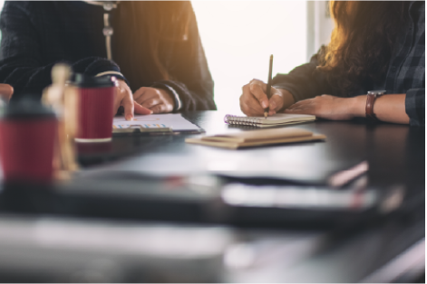 On voit un personne prenant des notes. Elle est accompagnés par d'autres personnes. Tous se trouvent autour d'un bureau avec des d'autres notes.